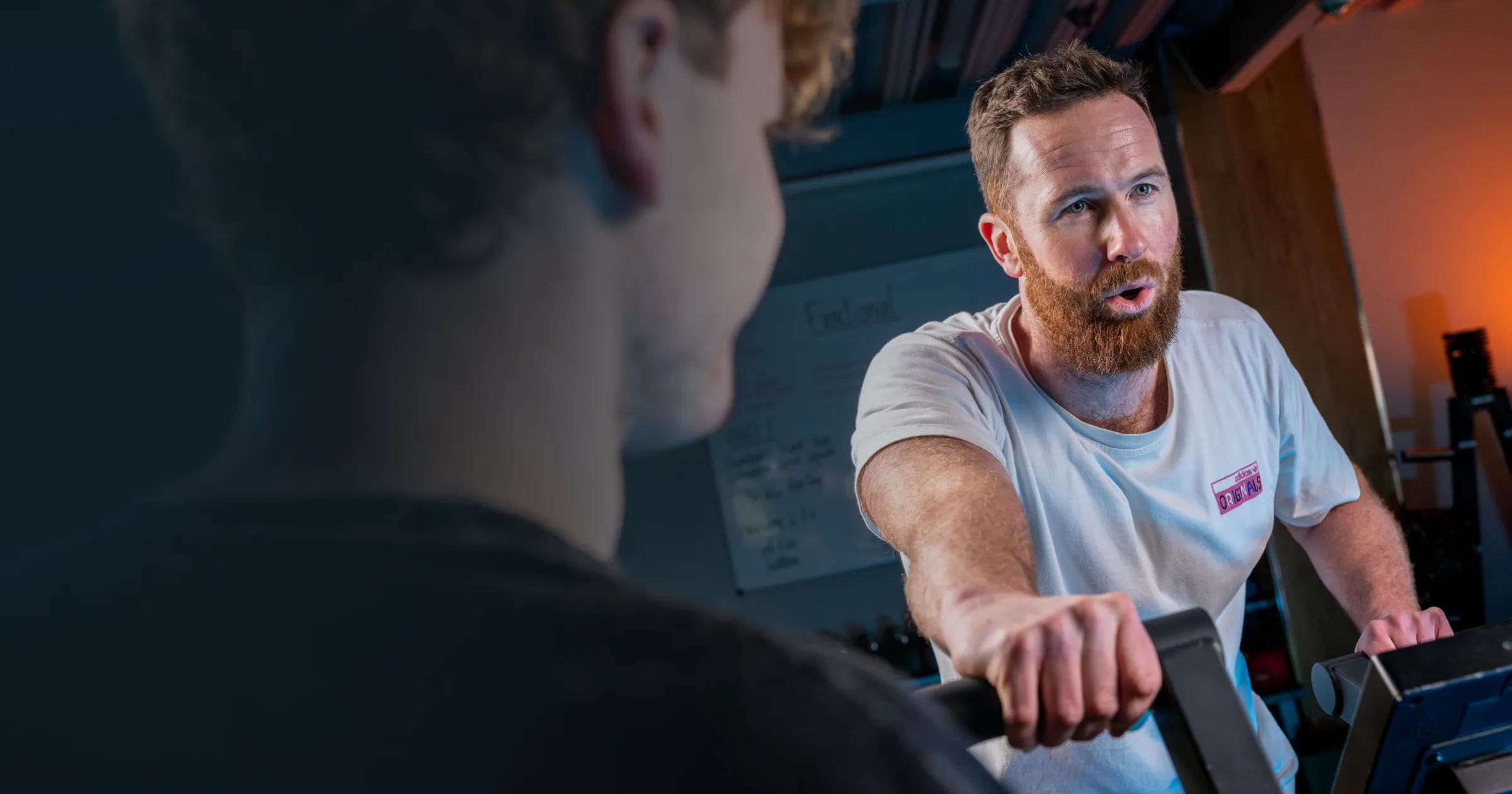 A focused trainer with a beard in a white t-shirt enthusiastically instructs a person on gym equipment. The setting is energetic and engaging.